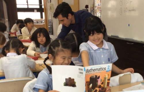 教室で外国人男性教師が立って生徒たちに授業をしている。画面手前の女の子二人は英語の教科書を開いて楽しそうに笑っている